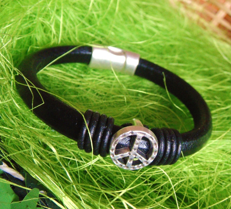 May include: Black leather bracelet with a silver peace sign charm and a magnetic clasp. The bracelet is set against a bright green background, highlighting the contrast between the dark leather and the shiny metal.