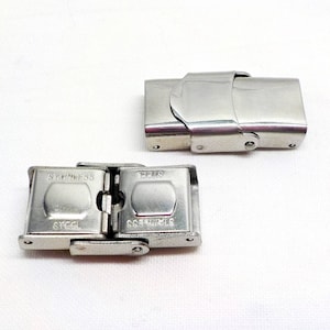 May include: Two silver metal watch band clasps. The clasp on the left is closed and the clasp on the right is open. The clasp on the left has the words "Stainless Steel" and "Steel" engraved on it.