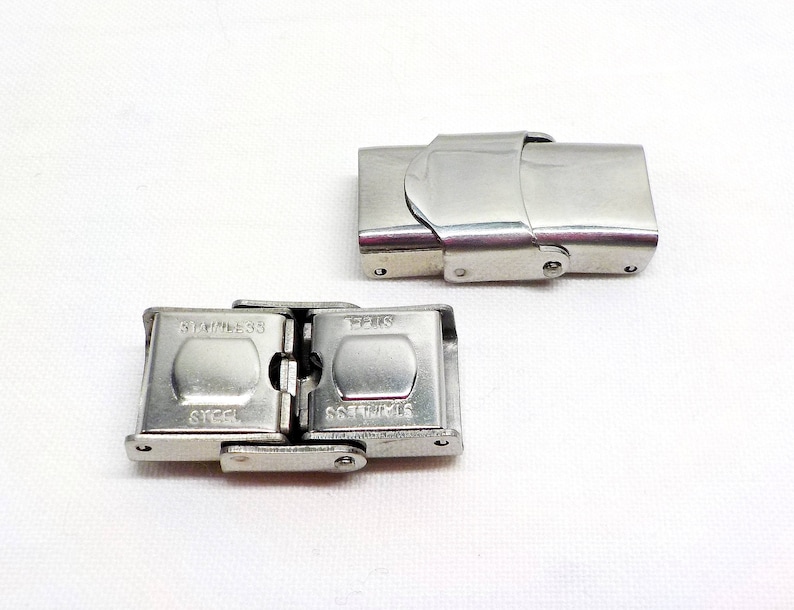 May include: Two silver metal watch band clasps. The clasp on the left is closed and the clasp on the right is open. The clasp on the left has the words "Stainless Steel" and "Steel" engraved on it.