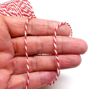 May include: Red and white baker's twine, a type of string used for wrapping and tying, is held between the fingers of a hand.