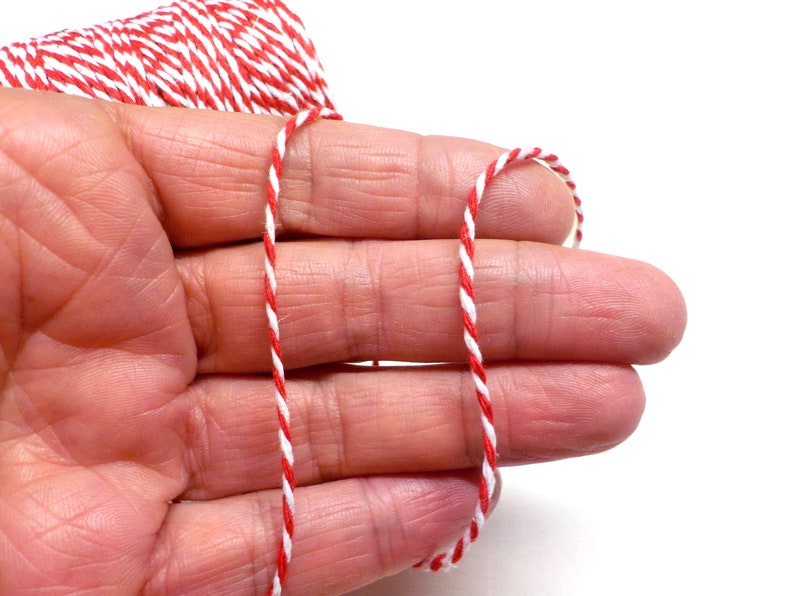 May include: Red and white baker's twine, a type of string used for wrapping and tying, is held between the fingers of a hand.