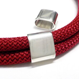 May include: Two silver metal cord ends for a red rope bracelet. The cord ends are rectangular with rounded edges.