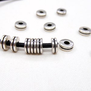 Op de afbeelding: Zilveren metalen kralen met een zwart leren koord. De kralen zijn rond en hebben een gegroefd ontwerp. Er zijn verschillende kralen verspreid rond het koord.