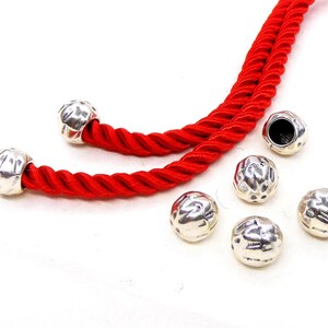 May include: Silver-colored, textured beads with a hammered appearance. The beads are shown with a bright red, twisted cord. The beads are round and have a dimpled surface. The cord is a vibrant red color.