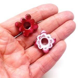 Puede incluir: Dos cuentas de metal de color rosa en forma de flor con un agujero en el centro. Las cuentas tienen un dise&ntilde;o blanco en los bordes.