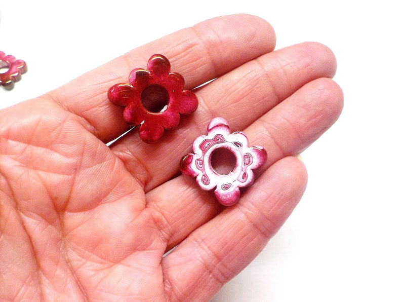 Puede incluir: Dos cuentas de metal de color rosa en forma de flor con un agujero en el centro. Las cuentas tienen un dise&ntilde;o blanco en los bordes.