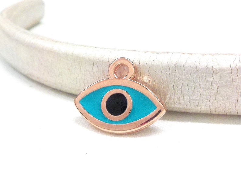 Puede incluir: Un amuleto de oro rosa en forma de ojo turco. El ojo es azul con una pupila negra y un contorno dorado.
