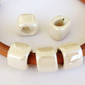 Peut inclure: Quatre perles en céramique blanche avec un trou au centre, enfilées sur un bracelet en cuir marron.
