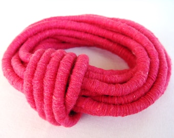 May include: A bright pink fabric bracelet made of tightly wound yarn. The bracelet is knotted in the front and has a circular shape.