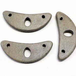 May include: Three gray ceramic crescent moon shapes with holes in the center. The shapes are slightly different sizes and have a rough texture.
