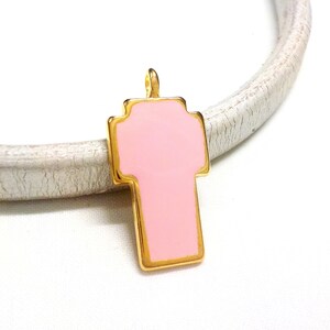 May include: A gold-toned cross charm with a pink enamel fill. The charm is on a white surface.