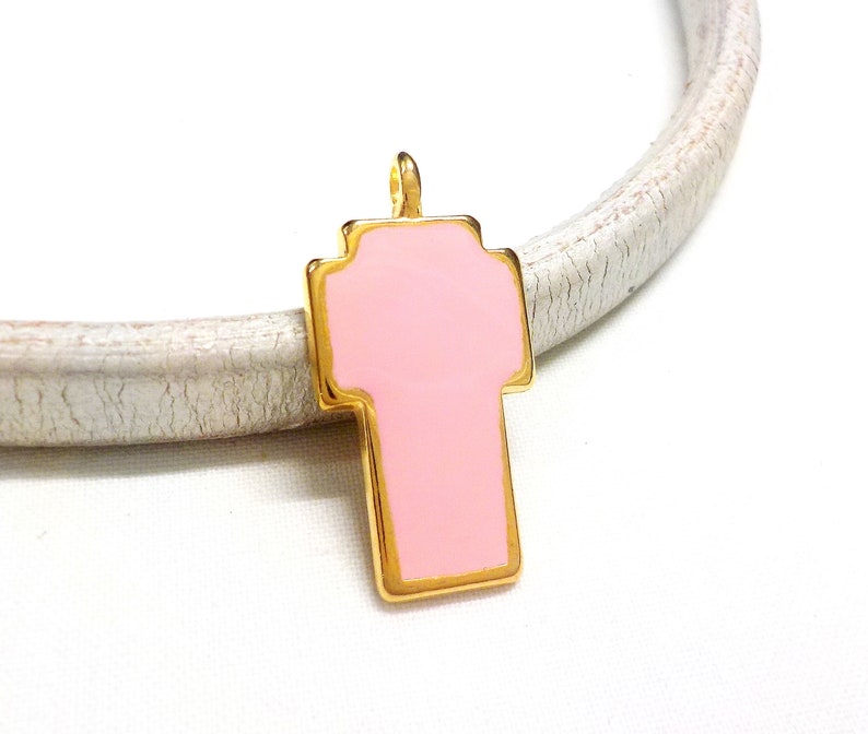 May include: A gold-toned cross charm with a pink enamel fill. The charm is on a white surface.