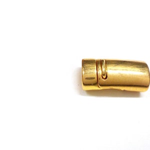 May include: A gold-colored magnetic clasp, likely for jewelry or accessories, lies on a white background. The clasp is rectangular with rounded edges and a small, hinged section. The metal has a slightly aged appearance.