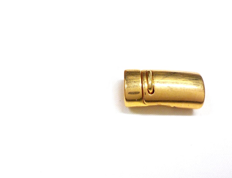 May include: A gold-colored magnetic clasp, likely for jewelry or accessories, lies on a white background. The clasp is rectangular with rounded edges and a small, hinged section. The metal has a slightly aged appearance.