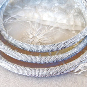 May include: Three silver-colored, mesh-like necklaces are arranged in a layered composition. The necklaces are circular and have a textured appearance. The background is a soft, white fabric with delicate lace details, enhancing the necklaces' shine.