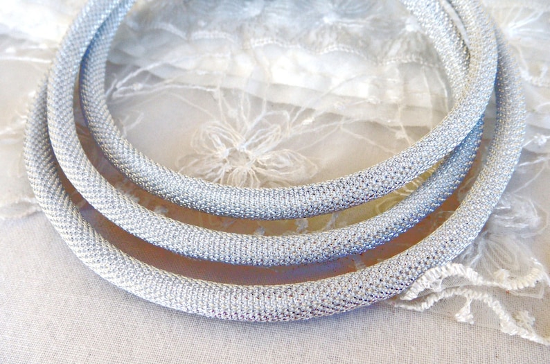 May include: Three silver-colored, mesh-like necklaces are arranged in a layered composition. The necklaces are circular and have a textured appearance. The background is a soft, white fabric with delicate lace details, enhancing the necklaces' shine.