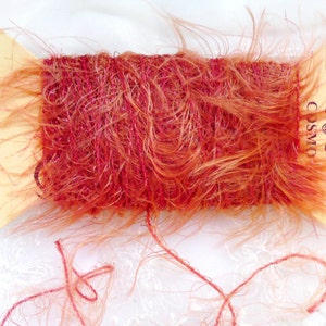 Puede incluir: Una bobina de hilo peludo rojo y naranja con la palabra "COSMO" impresa en la etiqueta.