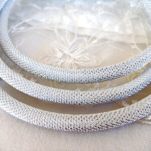 May include: Three silver-colored, mesh-textured necklaces are arranged in a curved pattern. The necklaces are made of a shiny, metallic material. The background is a soft, white fabric with a floral lace pattern.