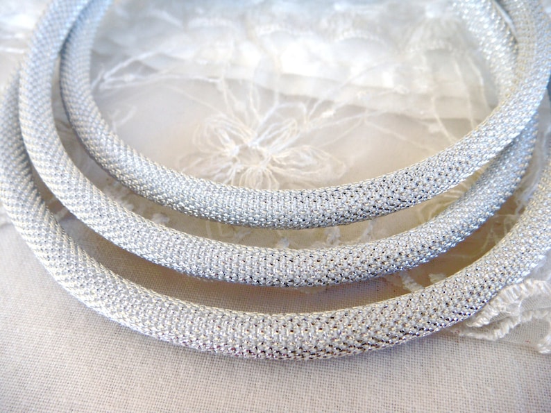 May include: Three silver-colored, mesh-textured necklaces are arranged in a curved pattern. The necklaces are made of a shiny, metallic material. The background is a soft, white fabric with a floral lace pattern.
