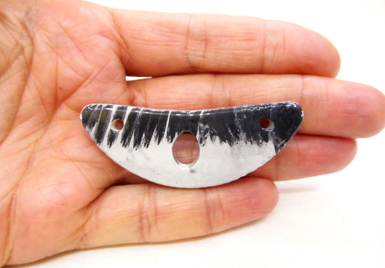 May include: A white and black ceramic pendant with a crescent shape and two holes. The pendant has a black and white striped pattern.