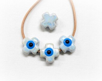 Puede incluir: Un collar azul claro con cuatro dijes de cerámica. Tres de los dijes tienen forma de cruz con un ojo turco azul en el centro. El cuarto dije es una cruz simple.
