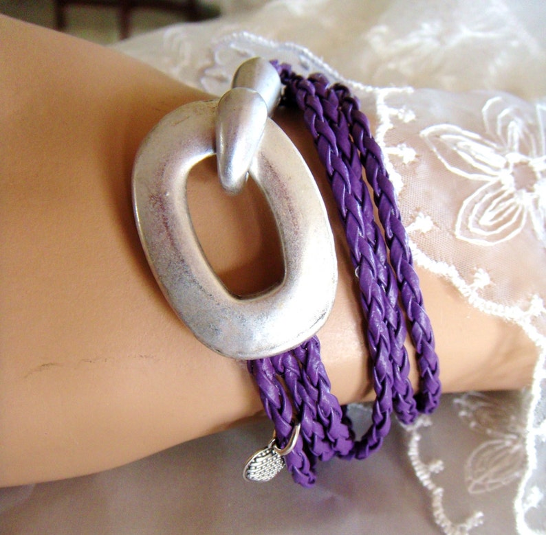 May include: A purple braided leather wrap bracelet with a silver oval charm. The charm has a large open circle in the center.