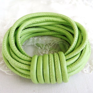 May include: A lime green leather bracelet with a stitched design. The bracelet is made of several looped strands of leather, held together by a thicker, rounded section. The bracelet is a bright, eye-catching accessory.