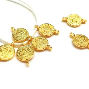 Op de afbeelding: Gouden metalen bedels met ingewikkelde ontwerpen, sommige met Arabische schrift, bevestigd aan een wit touwtje. De bedels zijn rond en hebben aan elke kant een lus om aan een ketting of armband te bevestigen.