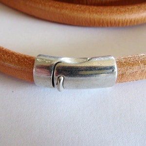 May include: A close-up of a tan leather bracelet with a silver-colored magnetic clasp. The bracelet is round and appears to be made of a single strand of leather. The clasp is rectangular and has a secure closure.
