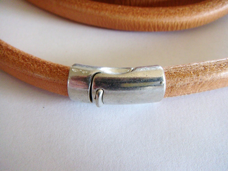 May include: A close-up of a tan leather bracelet with a silver-colored magnetic clasp. The bracelet is round and appears to be made of a single strand of leather. The clasp is rectangular and has a secure closure.
