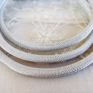 May include: Close-up of three silver-colored, textured, tubular necklaces. The necklaces are arranged in a circular pattern, with each one slightly overlapping the other. The texture appears to be a tightly woven mesh or knit, giving a subtle shimmer.