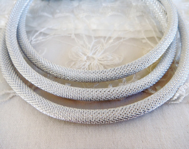 May include: Close-up of three silver-colored, textured, tubular necklaces. The necklaces are arranged in a circular pattern, with each one slightly overlapping the other. The texture appears to be a tightly woven mesh or knit, giving a subtle shimmer.