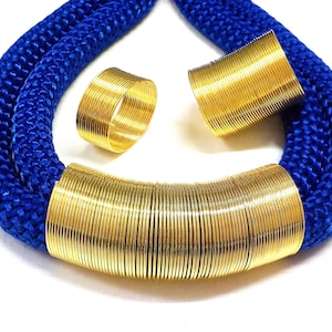 Puede incluir: Un collar de cuerda azul con un acento de metal dorado en forma de bobina gruesa. El collar se acompaña de dos anillos de metal dorado en forma de bobina más pequeños.