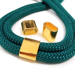 May include: Teal rope necklace with three gold-coloured metal clasps. The rope is thick and braided, and the clasps are rectangular and oval-shaped. The necklace is displayed on a white background.