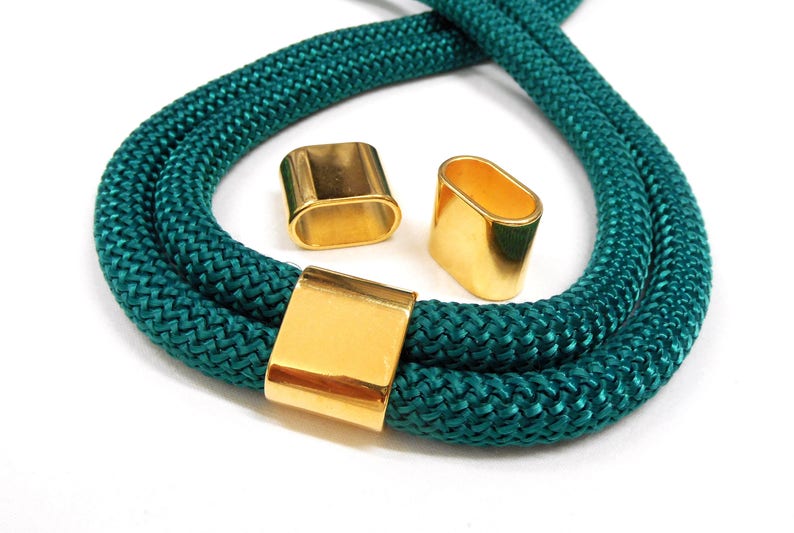 May include: Teal rope necklace with three gold-coloured metal clasps. The rope is thick and braided, and the clasps are rectangular and oval-shaped. The necklace is displayed on a white background.