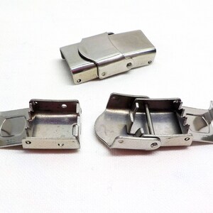 May include: Three silver metal watch band clasps. Two clasps are open and one is closed. The clasps are designed to be used with a watch strap.