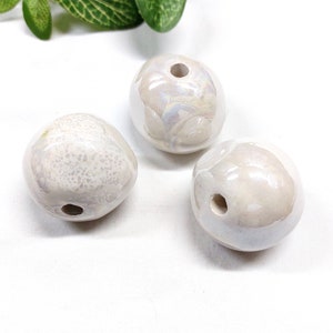 Könnte beinhalten: Drei weiße Keramikperlen mit einem Loch in der Mitte. Die Perlen haben eine perlmuttartige Oberfläche und sind leicht unregelmäßig geformt.