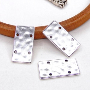 Puede incluir: Tres piezas de conexión metálicas rectangulares de color plateado con textura martillada y cuatro agujeros cada una. Las piezas están sobre una superficie de cuero marrón.