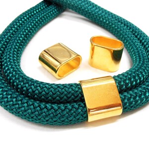 May include: Teal rope necklace with three gold-coloured metal clasps. The rope is thick and braided, and the clasps are rectangular and oval-shaped. The necklace is displayed on a white background.