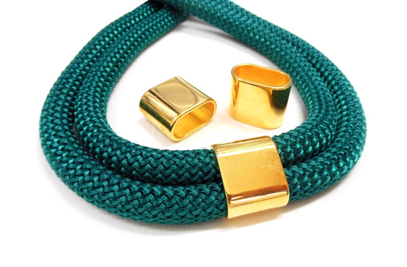 May include: Teal rope necklace with three gold-coloured metal clasps. The rope is thick and braided, and the clasps are rectangular and oval-shaped. The necklace is displayed on a white background.