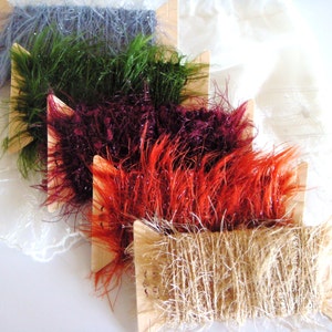 May include: Five spools of yarn in different colors and textures. The spools are arranged in a stack, with the top spool being a natural beige color, followed by a bright orange, a deep burgundy, a bright green, and a light blue spool.