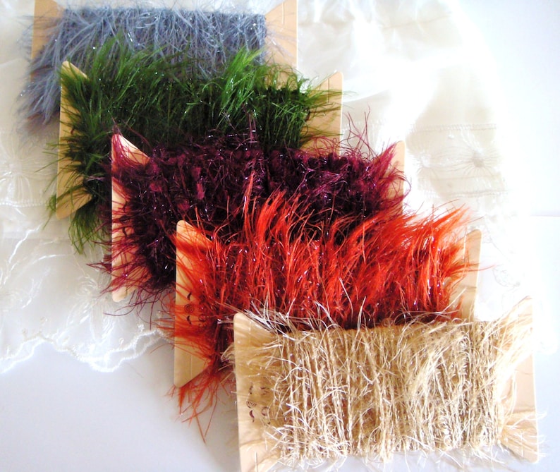 May include: Five spools of yarn in different colors and textures. The spools are arranged in a stack, with the top spool being a natural beige color, followed by a bright orange, a deep burgundy, a bright green, and a light blue spool.