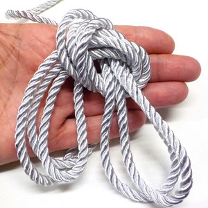 Puede incluir: Primer plano de una cuerda trenzada plateada, mostrando la textura y el detalle del cordón. La cuerda está enrollada y dispuesta en una mano, destacando su naturaleza suave y flexible.