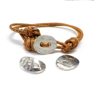 Può includere: Un braccialetto in corda marrone con chiusura a bottone argentata. Sono visibili anche due bottoni argentati aggiuntivi. Il braccialetto è annodato e sembra fatto a mano. Lo sfondo è bianco.