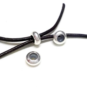 Pode incluir: Três pontas de cordão de metal prateado com um furo no centro. As pontas de cordão estão em um cordão de couro marrom.