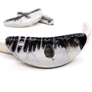 Peut inclure: Un pendentif en céramique blanche avec des coups de pinceau noirs et deux trous. Le pendentif a la forme d'un croissant de lune.