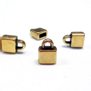 May include: Close-up of several small, square, brass-colored metal clasps with a loop on top. One clasp is attached to a piece of cork material. The clasps are on a white background.