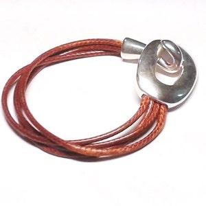 May include: A brown leather bracelet with a silver clasp. The clasp is a unique design with a large oval shape and a smaller oval cut out in the center.