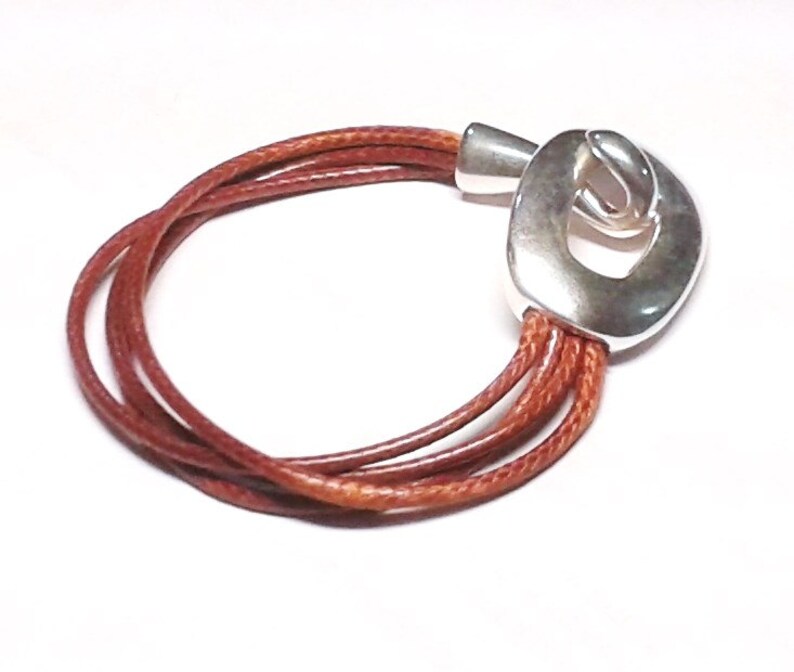 May include: A brown leather bracelet with a silver clasp. The clasp is a unique design with a large oval shape and a smaller oval cut out in the center.