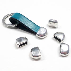 Könnte beinhalten: Ein türkisfarbener Leder-Schlüsselanhänger mit einem silbernen Schlüsselring und sechs silbernen Metall-Endkappen. Die Endkappen sind in Form von kleinen, abgerundeten Rechtecken.